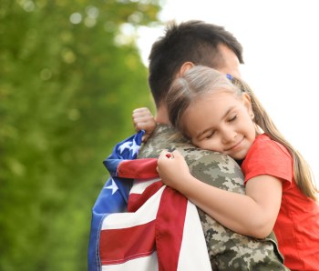 Father in military uniform with American flag hugging his little daughter at green park