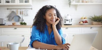 Indoor shot of happy young Afro American woman using portable computer and smiling, enjoying modern technology while paying bills for rent, gas and electricity online, sitting at kitchen table