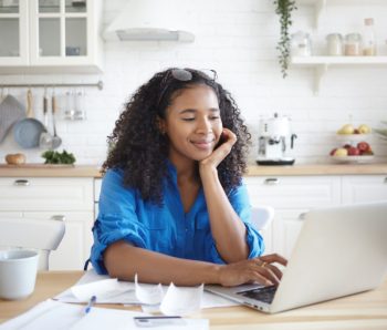 Indoor shot of happy young Afro American woman using portable computer and smiling, enjoying modern technology while paying bills for rent, gas and electricity online, sitting at kitchen table