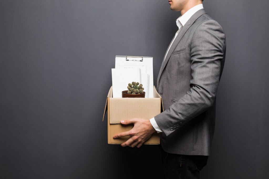 Close up of fired man employee hiding behind box with personal items on grey background