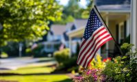 American flag waving in front of residential homes with green lawns and colorful flowers, symbolizing patriotism and community in suburban neighborhoods.