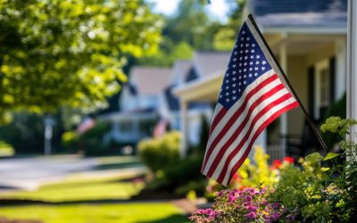 American flag waving in front of residential homes with green lawns and colorful flowers, symbolizing patriotism and community in suburban neighborhoods.
