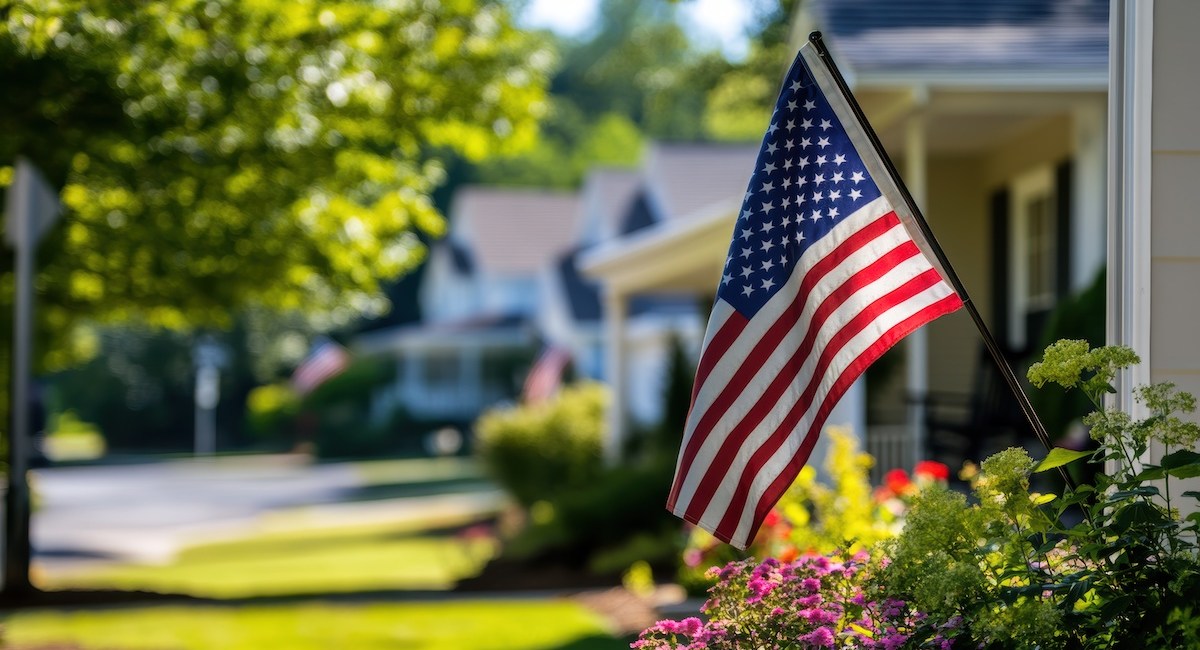 American flag waving in front of residential homes with green lawns and colorful flowers, symbolizing patriotism and community in suburban neighborhoods.