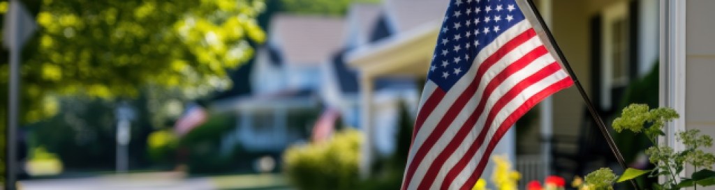 American flag waving in front of residential homes with green lawns and colorful flowers, symbolizing patriotism and community in suburban neighborhoods.